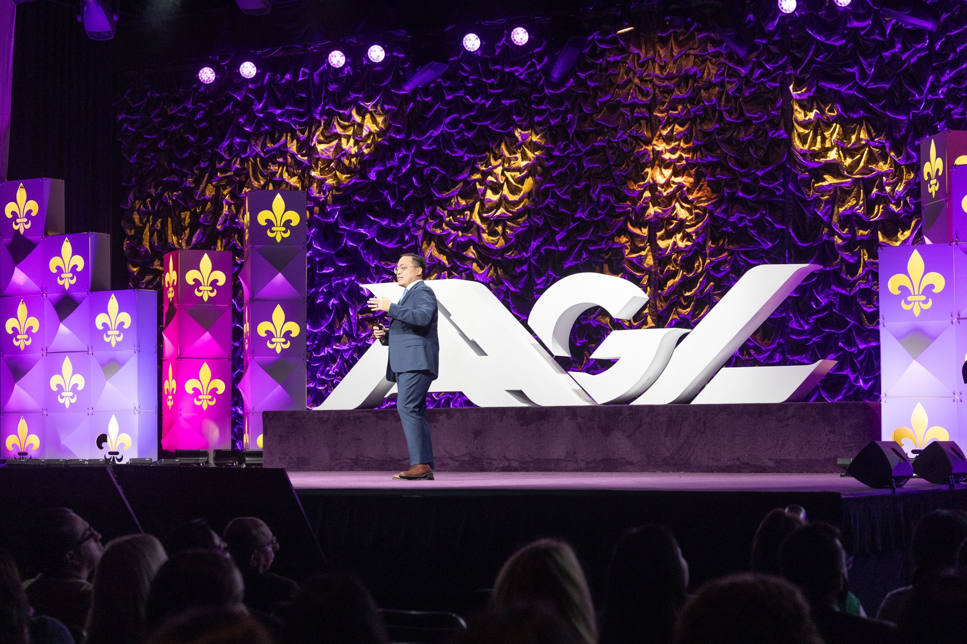 AAGL Global Congress main stage — speaker presenting in front of giant 3D AAGL letters with Mardi Gras themed purple and gold stage design